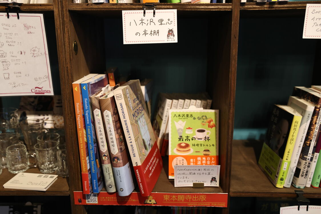 Notre interview s’est déroulée à la librairie partagée (share bookstore) « Neko no tana » du quartier de Jinbôchô. Chaque étagère a son propre patron, qui ouvre là une mini-librairie où il présente ses livres préférés. Yagisawa y a sa propre étagère où se trouvent les éditions en langue étrangères de ses romans.