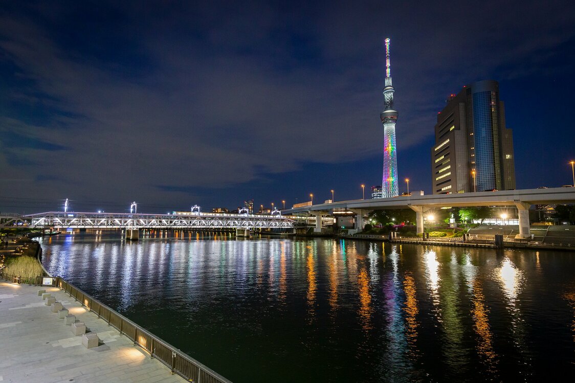 Tras la puesta de sol, durante una hora, destaca la hermosa gama de colores con una base blanca de gran simplicidad. En la fotografía también se observa, en la parte inferior del puente ferroviario de Sumidagawa, el paso peatonal Sumida River Walk que conecta la zona con Asakusa y, a la derecha de la torre, el edificio del ayuntamiento del distrito de Sumida.