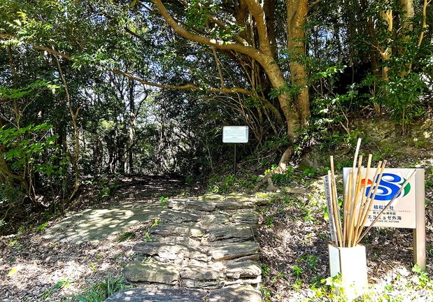 The entrance to Karematsu Jinja. From here, a five-minute walk up a stone stairway leads to the main hall. There are walking sticks for pilgrims in front of the sign. (© Amano Hisaki)