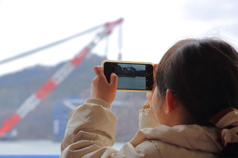 A young schoolgirl from the island photographs the new bridge pillars during a tour of the site for Ōshima residents. Her worried question to her father, “Is the ferry really going to disappear?” hints at the uncertainties that remain in the islanders’ future.