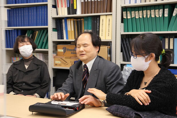 Fukushima demonstrates the use of his BrailleSense device by reading out the news from an online newspaper. Sitting to his left is finger braille interpreter Maeda Atsumi. (© Hanai Tomoko)