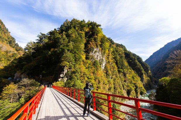 The vermillion-lacquered Okukane Bridge on the way to Babadani Onsen adds a spot of color to the lush greenery. In the background to the right is Kurobe River Power Station No. 3.