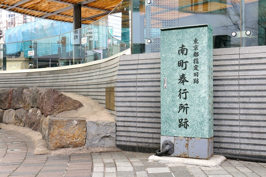 La stèle marquant l’emplacement de l’ancien poste de magistrature du Sud se dresse sur la place devant la gare JR de Yûrakuchô. Sur la gauche, on peut voir l’alignement de pierres qui bordait autrefois le caniveau, aujourd’hui exposé comme monument commémoratif. (Pixta)