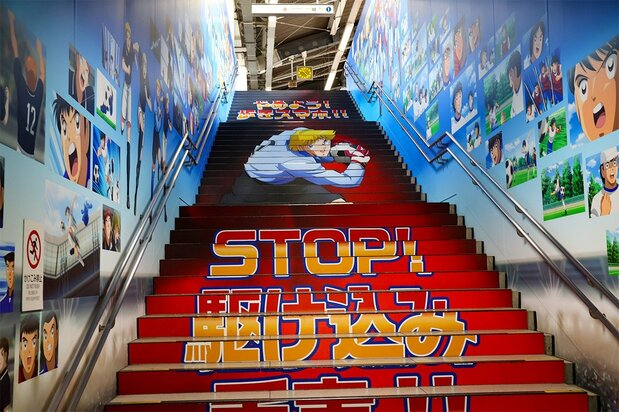 Un escalier vers le quai de la gare de Yotsugi. Les murs présentent de nombreux dessins des personnages de Captain Tsubasa. 