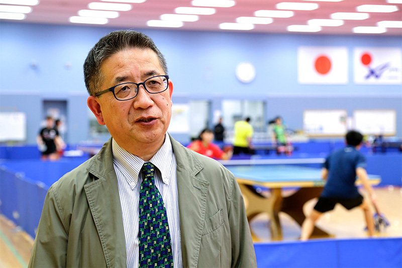 味の素ナショナルトレーニングセンターで取材に答える前原正浩氏。後方は、練習するJOCエリートアカデミーの選手たち。撮影＝長坂 芳樹