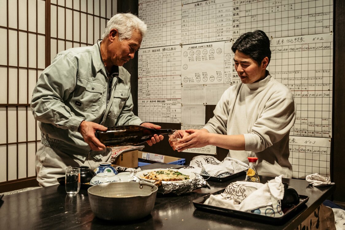 夜ごはんは蔵の酒とともに。杜氏を務める市川直道さん（44・右）に、ベテランの滝沢善治さん（69）が酒を注ぐ。午後6時には食べ終えて、明日に備える。壁には工程表が張ってある