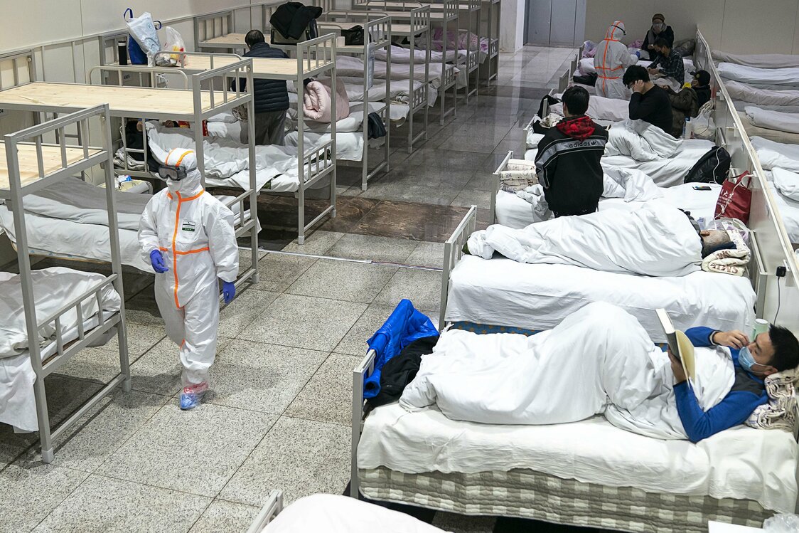 Медицинский работник в защитном костюме проходит мимо пациентов с коронавирусом, которые проходят лечение во временной больнице, расположенной в выставочном центре в Ухане, провинция Хубэй, 5 февраля 2020 г. (© Chinatopix / AP / Aflo)