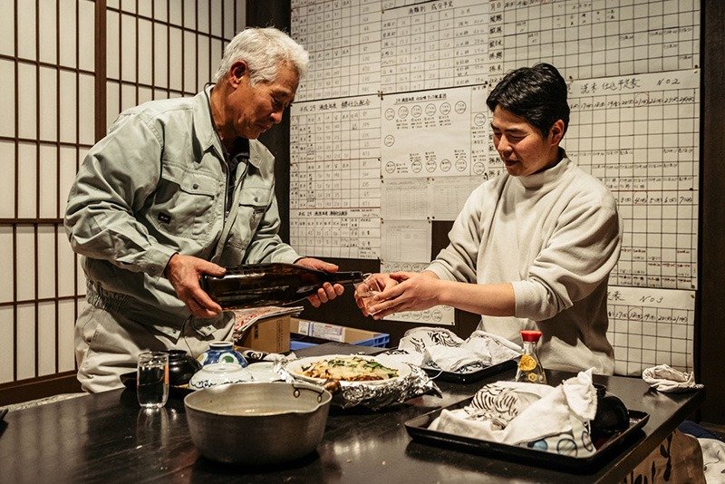 La cena se sirve acompañada de sake. A la derecha, el maestro jefe de la bodega, Ichikawa Naomichi, de 44 años. Le sirve todo un veterano de la profesión, Takizawa Zenji, de 69 años. Cuando terminan de cenar, a las 18:00, comienzan los preparativos para la siguiente jornada. En la pared, los horarios de trabajo.