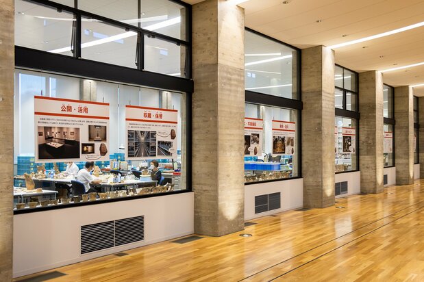 Staff restore artifacts inside the glass-walled workroom. Panels explain the workflow.