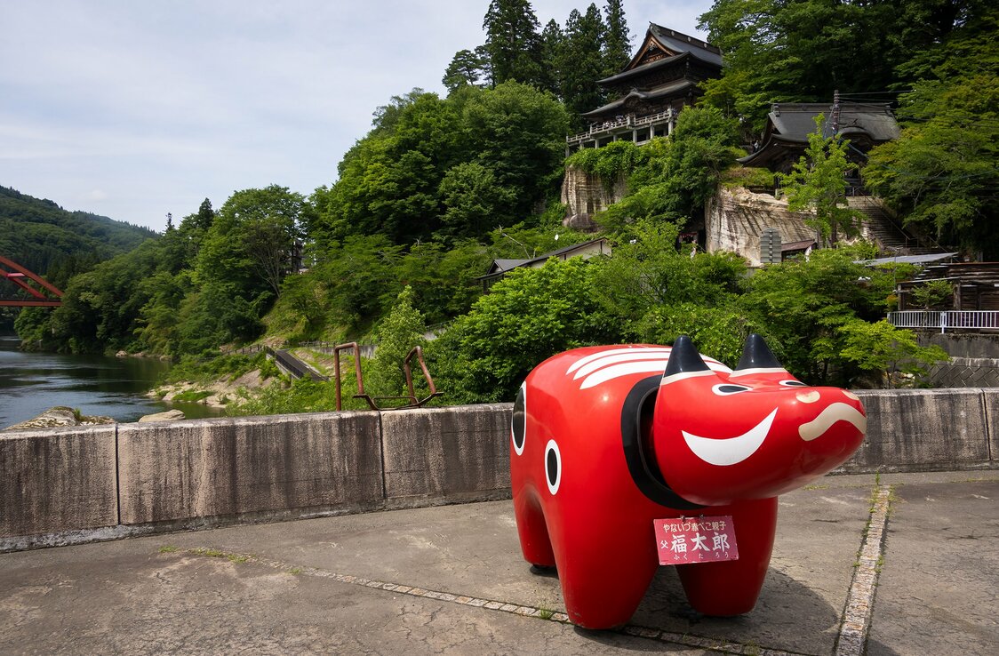 At Kiyohime Park in Yanaizu, visitors can pose with Fukutarō, a giant akabeko. Legend has it that red cows carried timber from the Tadami River, seen at left, up the cliff to the temple hall above. (© Nippon.com)