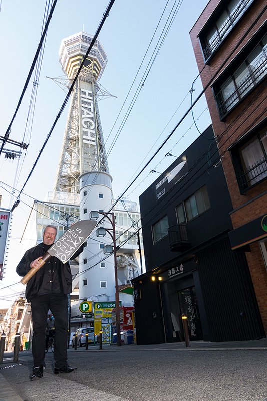 Улыбающийся Вийон Хайберг на фоне магазина Tower Knives Osaka и башни Цутэнкаку