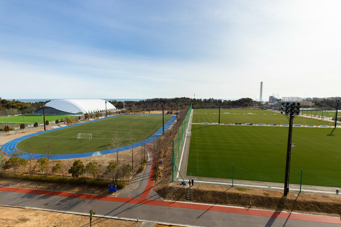 Les terrains vus de la salle panoramique du centre d’entraînement ; à droite, trois terrains en gazon naturel, un terrain synthétique entouré d’une piste d’athlétisme ; au fond à gauche, le terrain d’entraînement couvert et derrière, s’étend l’océan Pacifique.