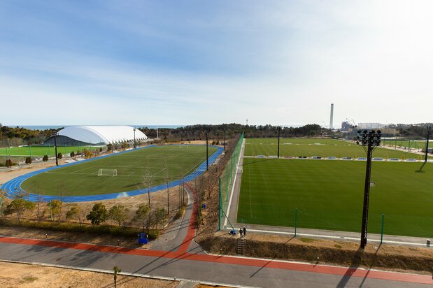 Los campos deportivos del J-Village desde su Edificio Central. Los tres campos que ocupan la mitad derecha de la fotografía son de césped natural. El espacio interior de la pista de atletismo de la izquierda, de césped artificial. Por encima de la carpa puede divisarse, al fondo, el océano Pacífico.