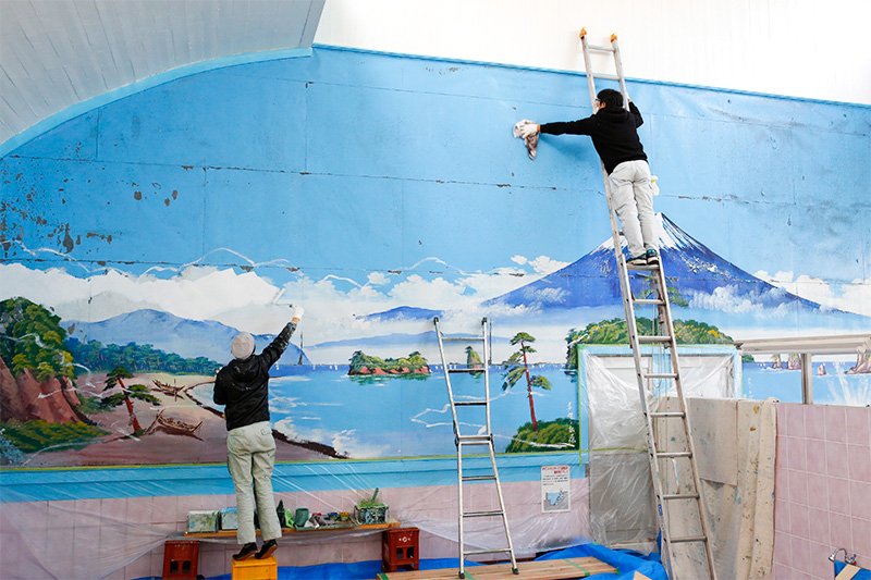 Tras eliminar las partes desconchadas, Tanaka comienza a pintar con el azul cielo. Sobre la escalera podemos ver a Komamura Yoshikazu, el marido y socio de la pintora.