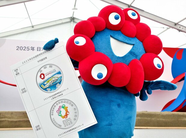 手持大阪关西世博会纪念币宣传板的世博会吉祥物“脉脉”（时事社）