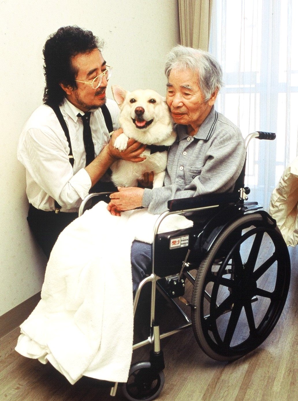 「待っていたよ」とチロリに触れる長谷川外吉さん（右）。チロリに励まされてリハビリに努め、要介護5の状態から自力で立てるまで回復を見せた（左は大木トオルさん）　写真提供＝国際セラピードッグ協会