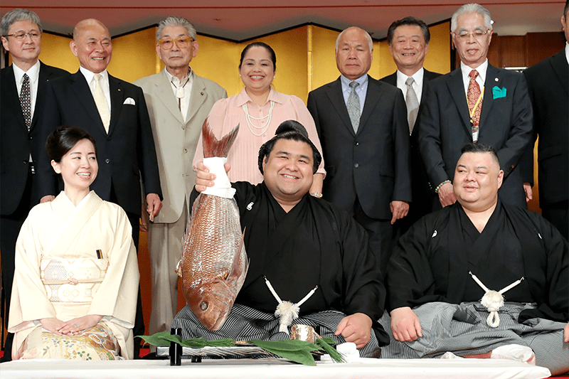 Takayasu (delante, en el centro) sostiene un tai (besugo) después de la ceremonia formal en la que se le comunica su ascenso en Tokio el 31 de mayo de 2017. Takanotsuru (sentado a la derecha) es el líder de su dōjō Tagonoura. Su padre Eiji (en el fondo, el tercero por la derecha) y su madre filipina Bebelita (detrás, en el centro) también están presentes. (© Jiji)