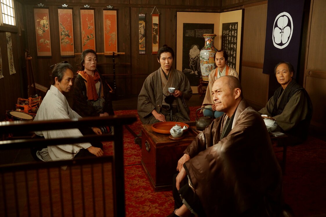 Sōichirō recopila los relatos de los miembros de la compañía Morita-za, entre ellos el autor y director de la obra, Shinoda Kinji (Watanabe Ken, al frente), y descubre la sorprendente verdad que se esconde tras la venganza. (© 2026 Comité de Producción de Kobikichō no adauchi © 2023 Nagai Sayako / Shinchōsha)