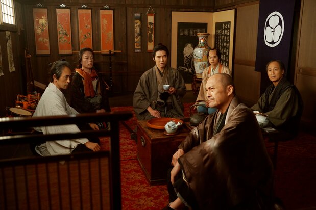 Sōichirō recopila los relatos de los miembros de la compañía Morita-za, entre ellos el autor y director de la obra, Shinoda Kinji (Watanabe Ken, al frente), y descubre la sorprendente verdad que se esconde tras la venganza. (© 2026 Comité de Producción de Kobikichō no adauchi © 2023 Nagai Sayako / Shinchōsha)