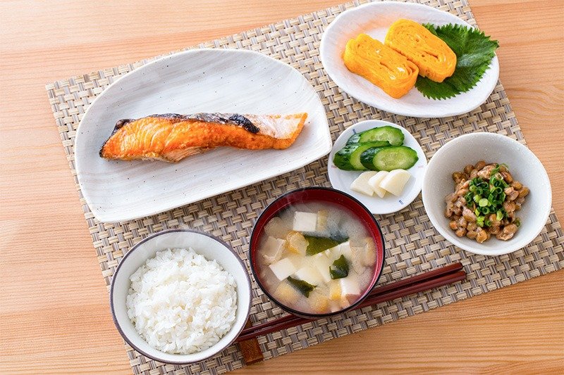 Desayuno tradicional japonés. Desde abajo a la derecha y siguiendo el sentido de las agujas del reloj: nattō con cebolleta o negi, sopa de miso con tofu y alga wakame, arroz cocido, salmón a la sal, encurtidos y tamagoyaki.