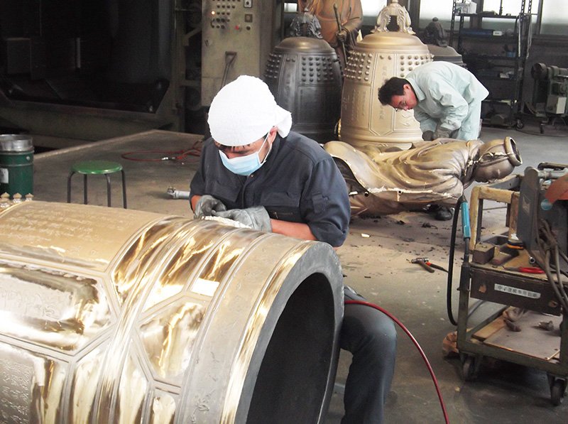 Des artisans polissant à la main une cloche qui vient de sortir du moule. A l’arrière plan, on voit un statue de bouddha en fabrication.