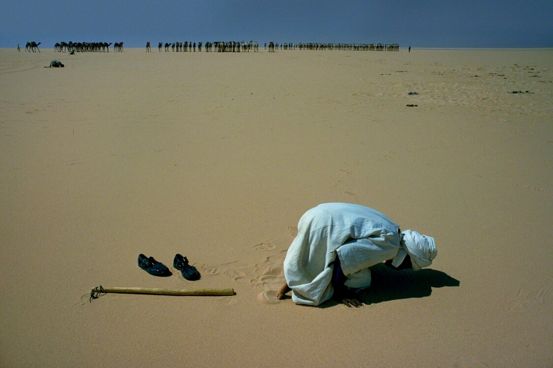 Après un déjeuner rapide, l’homme s’éloigne du groupe, s’immerge dans l’étendue de sable brûlant du désert, et prie en direction de la Mecque. Sa prière achevée, il retourne à l’arrière de la caravane. Photo prise en 1975 à Fezzan, dans le sud-ouest de la Libye. (Extrait de l’album « Sahara, entre ciel et sable »)